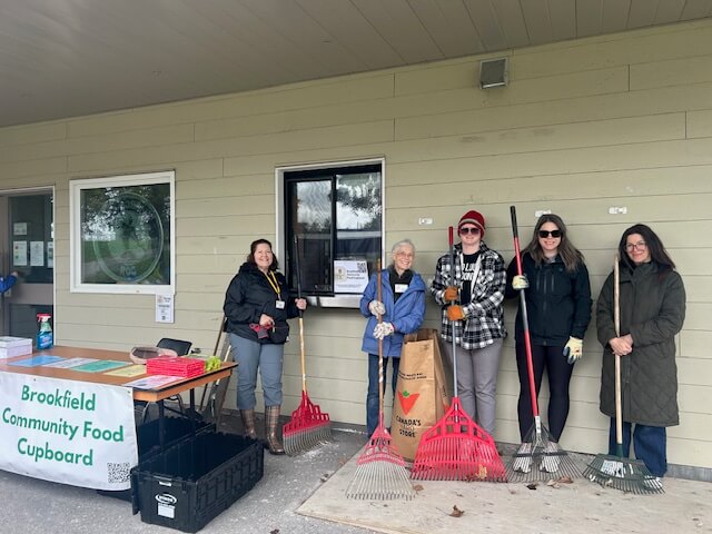 Five volunteers stand outside the Brookfield Community Food Cupboard holding rakes and brooms, with a registration table and supplies visible to the left.