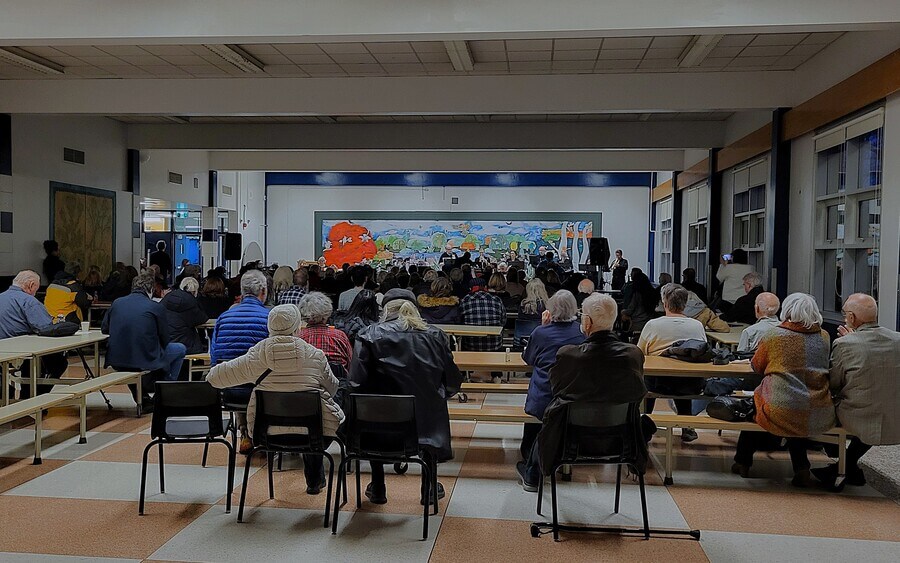 Audience members sit at tables facing a stage with a vibrant, colourful mural in a community hall with checkered floors and large windows.