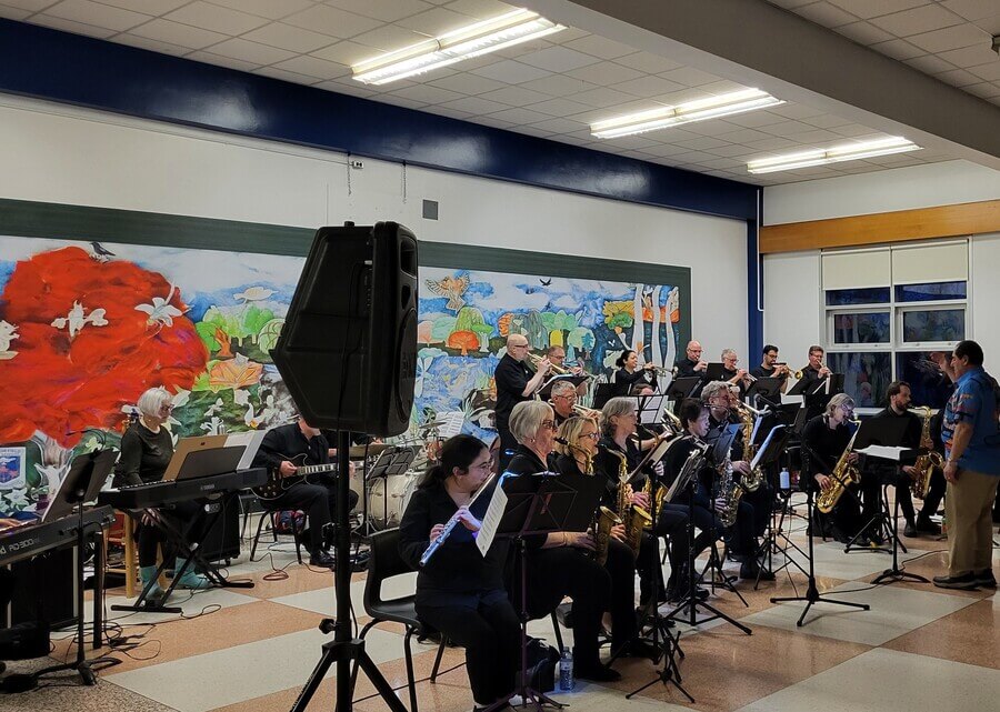 A community band performs with saxophones, flutes, keyboards and brass instruments in front of a vibrant mural featuring red poppies, in a hall with blue trim and checkered floors.