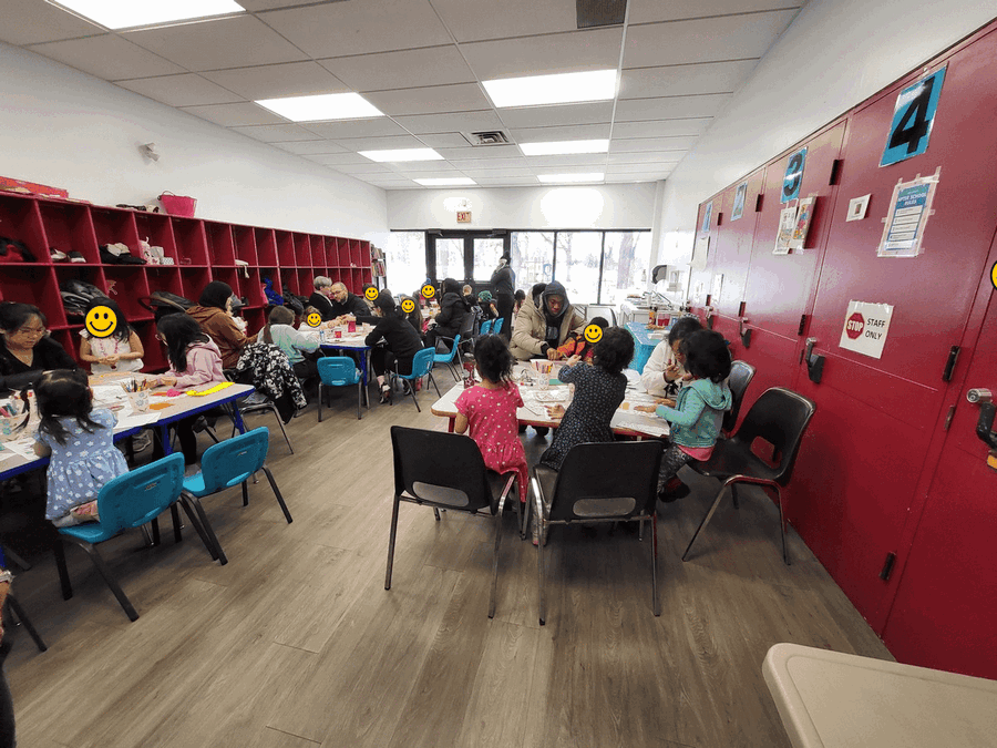 "A bright community room with red walls and lockers where children and adults sit at tables with blue and black chairs, participating in a spring-themed crafts and colouring event. Participants engage in creative activities together in this welcoming, inclusive community space with wood-look flooring and large windows.